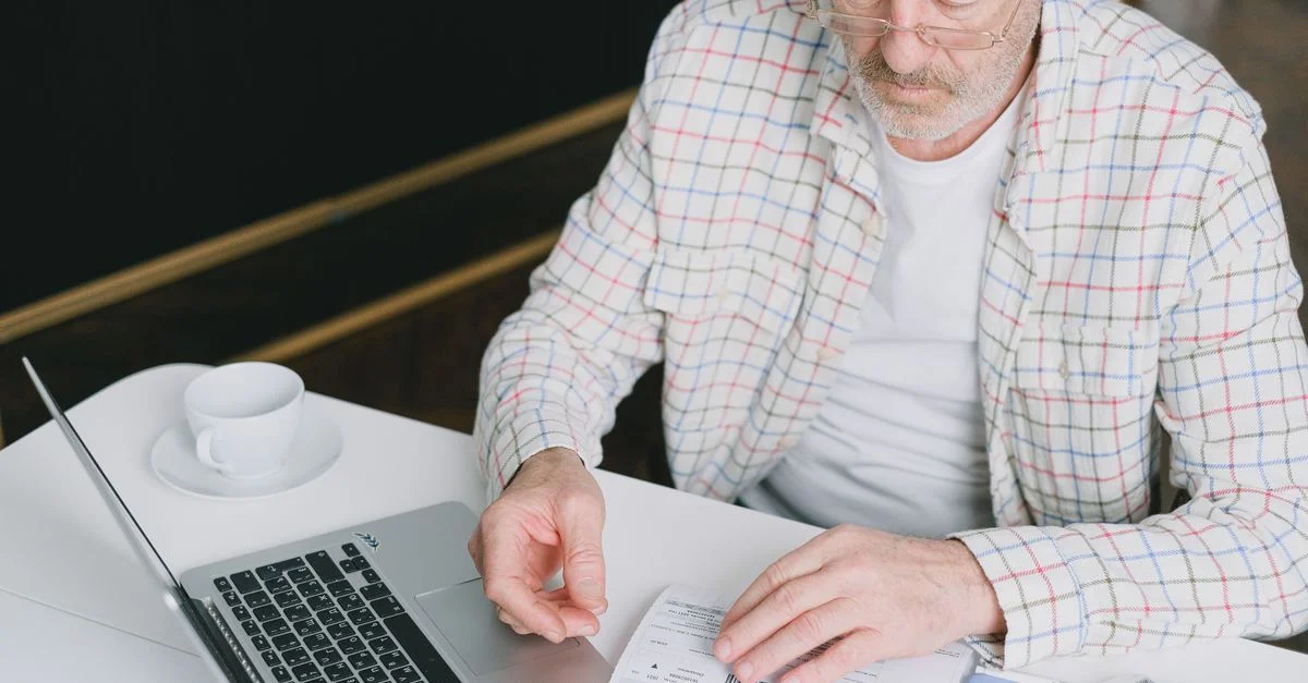 Homme âgé avec des lunettes révisant des documents sur un ordinateur portable. Cadre intérieur avec lumière naturelle.